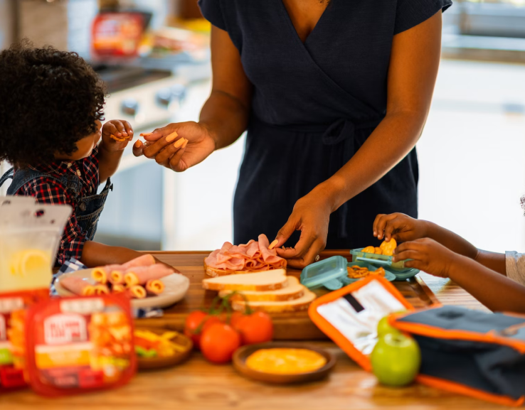 A woman preparing sandwiches in the kitchen with two young children helping and tasting the food