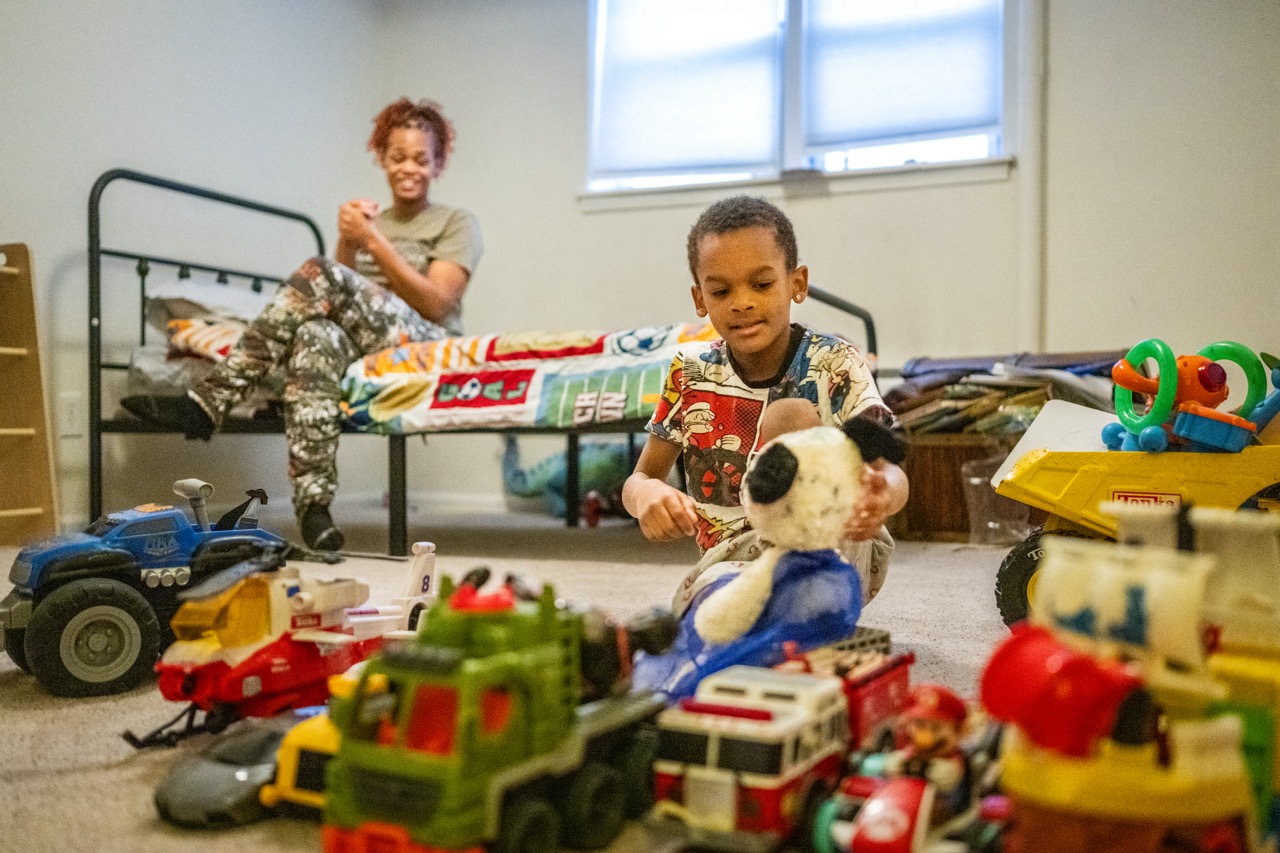 a young boy plays with toys, with his mother in the background