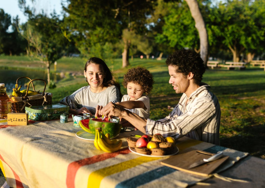 A family of three enjoying a picnic outdoors with various fruits and food on the table.